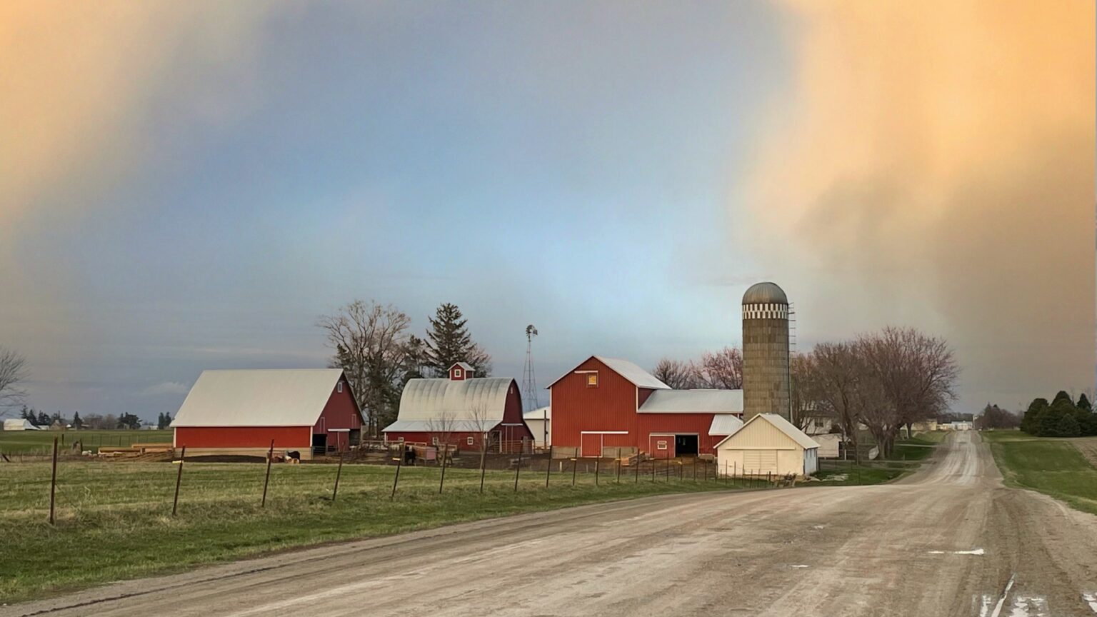 A rural farm scene with red barns, a silo, and a dirt road under a cloudy sky with orange and blue hues.