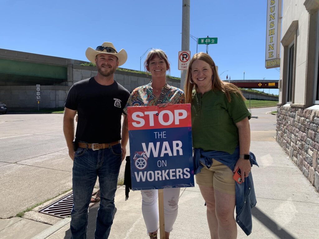 Three people stand on a sidewalk holding a sign that says "STOP THE WAR ON WORKERS". The person on the left wears a cowboy hat and a black t-shirt, the person in the middle wears a colorful patterned shirt, and the person on the right wears a green shirt and khaki shorts. A street sign for W 4th St is visible in the background, as well as a sign for Sunshine Restaurant.