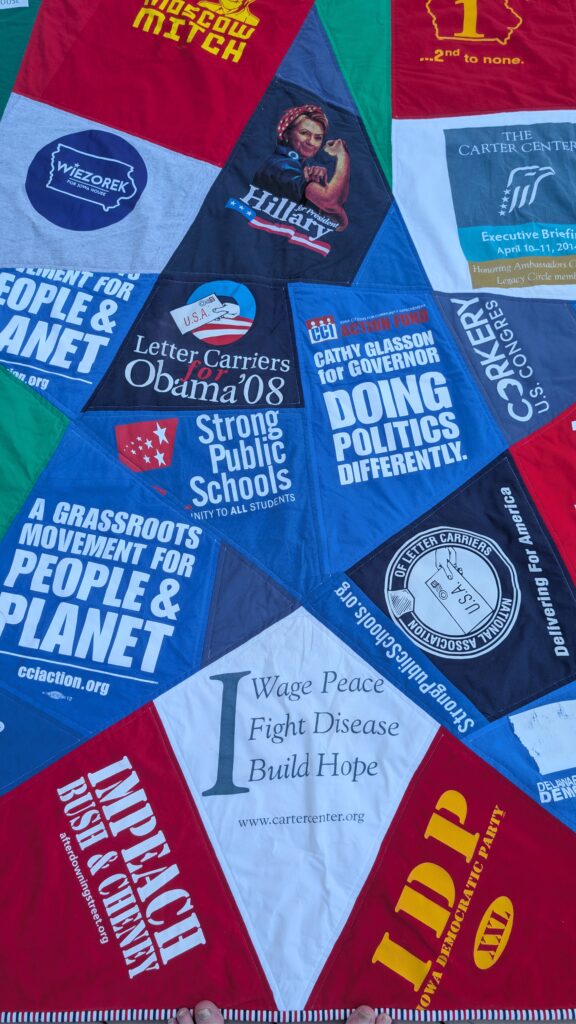 A political quilt featuring various campaign slogans and logos, including "Wiezorek for Iowa House," "Hillary for President," "Letter Carriers for Obama '08," "Strong Public Schools," "A Grassroots Movement for People & Planet," "The Carter Center," "Corkery U.S. Congress," "Impeach Bush & Cheney," "Iowa Democratic Party," and "Wage Peace, Fight Disease, Build Hope.