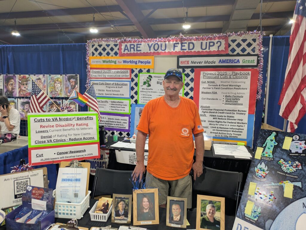 Man in an orange t-shirt stands at a political rally booth with signs about "Project 2025 - Playbook Proposed Cuts & Closings" and "Cuts to VA Medical Benefits.