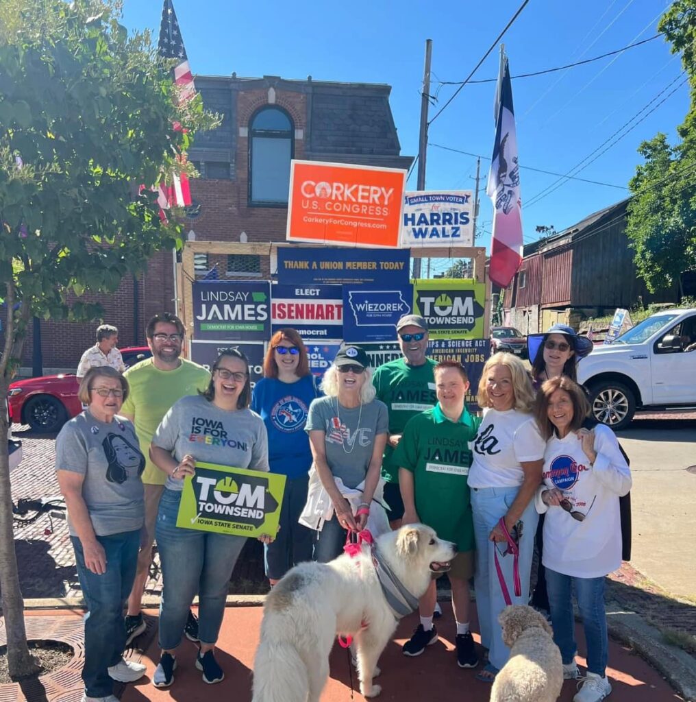 A group of people and two dogs stand in front of campaign signs for various candidates, including "CORKERY U.S. CONGRESS," "HARRIS WALZ," "LINDSAY JAMES," "ISENHART," "WIEZOREK," and "TOM TOWNSEND.