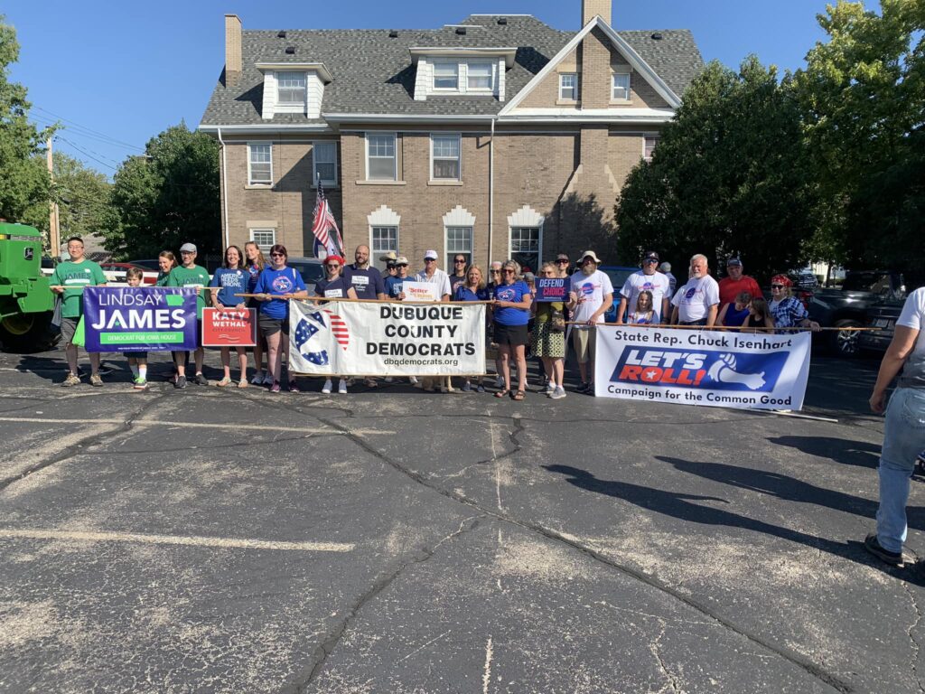 A group of people hold political banners in front of a brick building. The banners read "LINDSAY JAMES DEMOCRAT FOR IOWA HOUSE", "KATY WETHAL CITY COUNCIL", "DUBUQUE COUNTY DEMOCRATS dbqdemocrats.org", and "LET'S ROLL! Campaign for the Common Good".