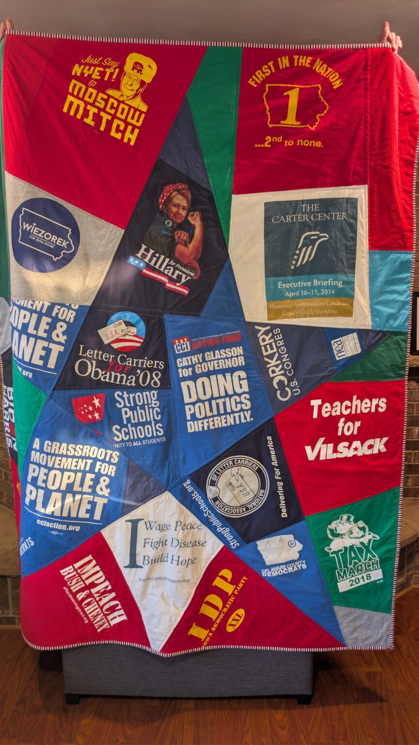 A colorful, politically themed quilt is displayed, featuring various campaign slogans and logos. The quilt is made of fabric panels sewn together, with prominent text and images. Some of the visible text includes "Just Say NYET! to MOSCOW MITCH," "FIRST IN THE NATION 1 ...2nd to none," "WIEZOREK FOR IOWA HOUSE," "Hillary for President," "THE CARTER CENTER," "Teachers for VILSACK," "A GRASSROOT