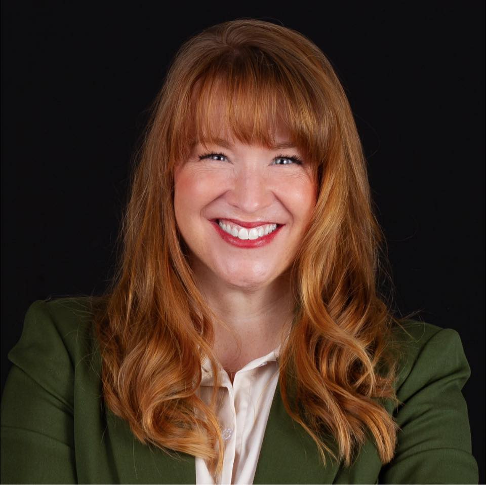 A woman with red hair and bangs smiles widely, wearing a green blazer over a cream-colored shirt.