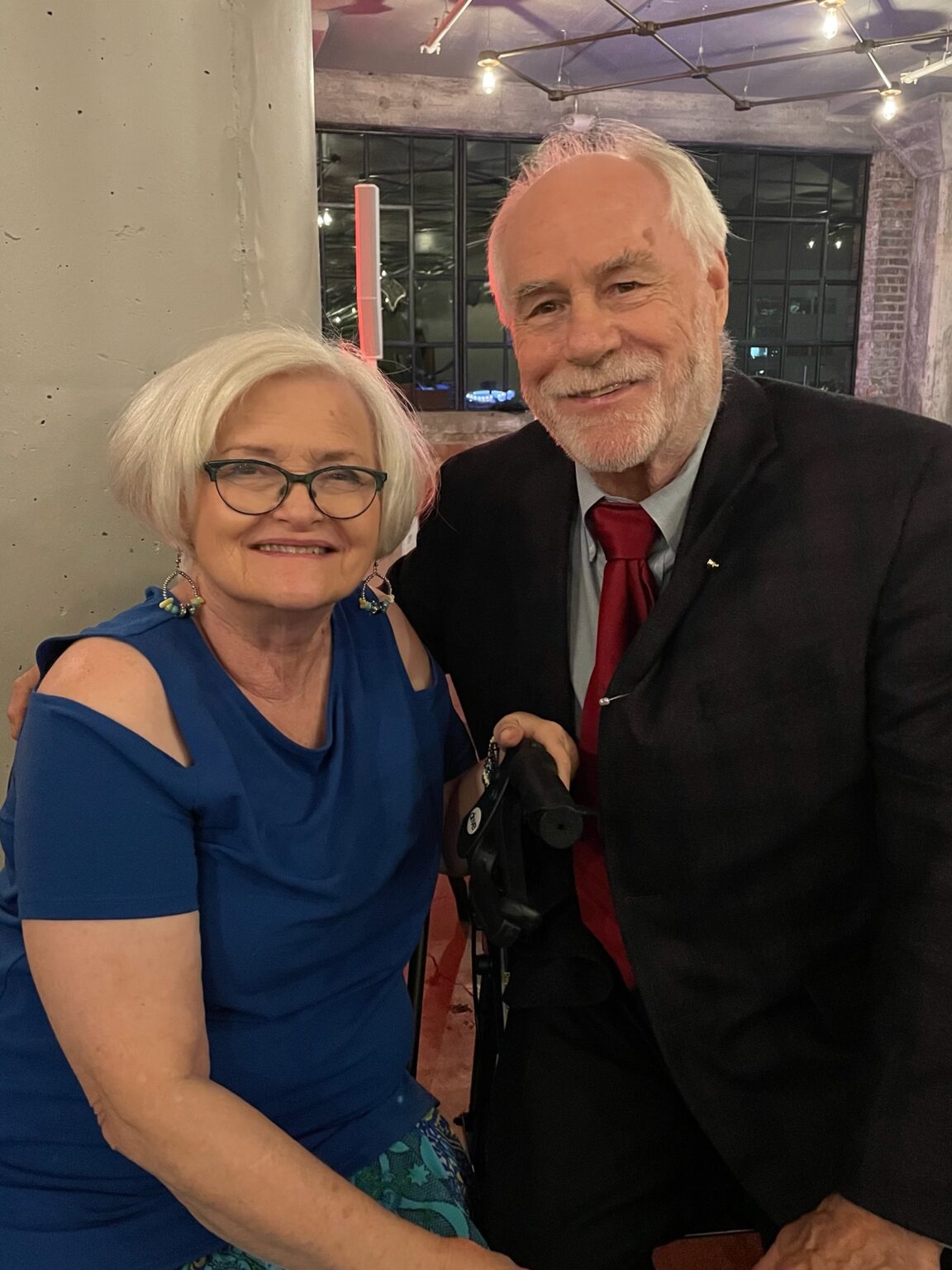 A smiling older couple poses for a photo. The woman on the left has short white hair, wears glasses, and a blue off-the-shoulder top. The man on the right has white hair and a beard, wears a black suit with a red tie, and holds a cane.