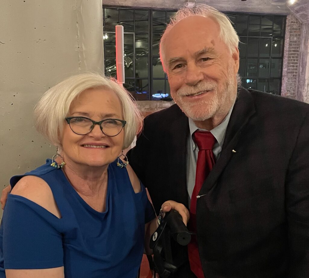 A smiling older couple poses for a photo. The woman on the left has short white hair, wears glasses, and a blue off-the-shoulder top. The man on the right has white hair and a beard, wears a black suit with a red tie, and holds a cane.