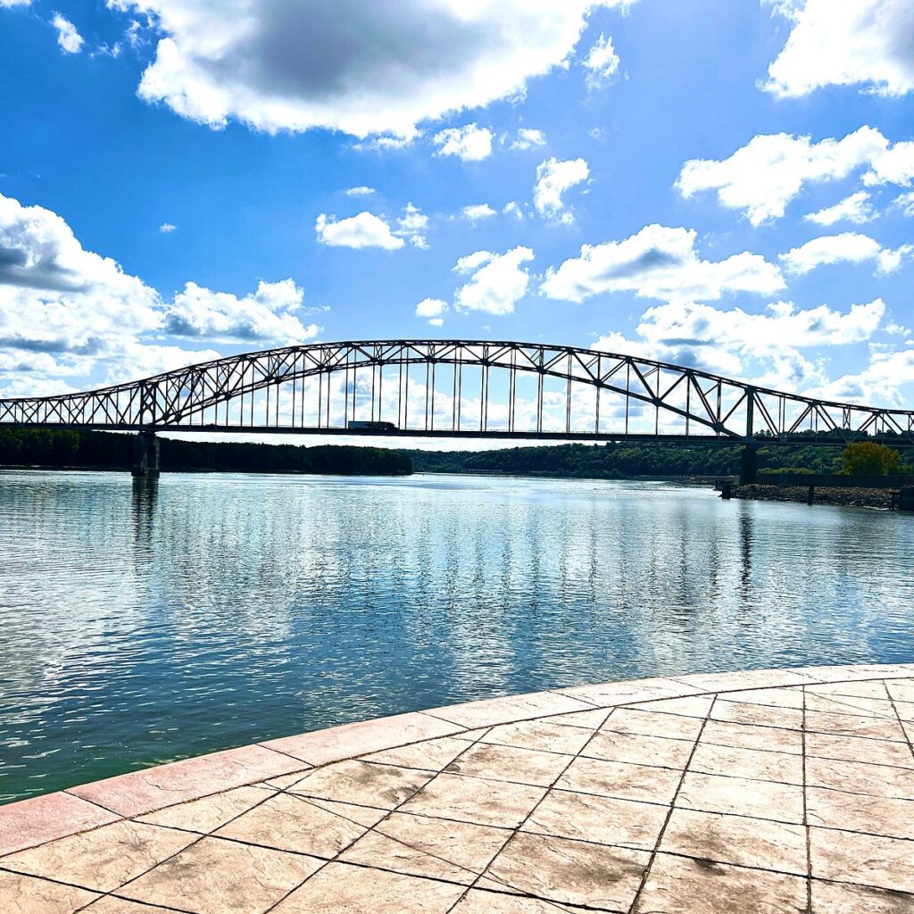 A wide, black steel truss bridge spans a calm, blue river under a bright sky with fluffy white clouds.