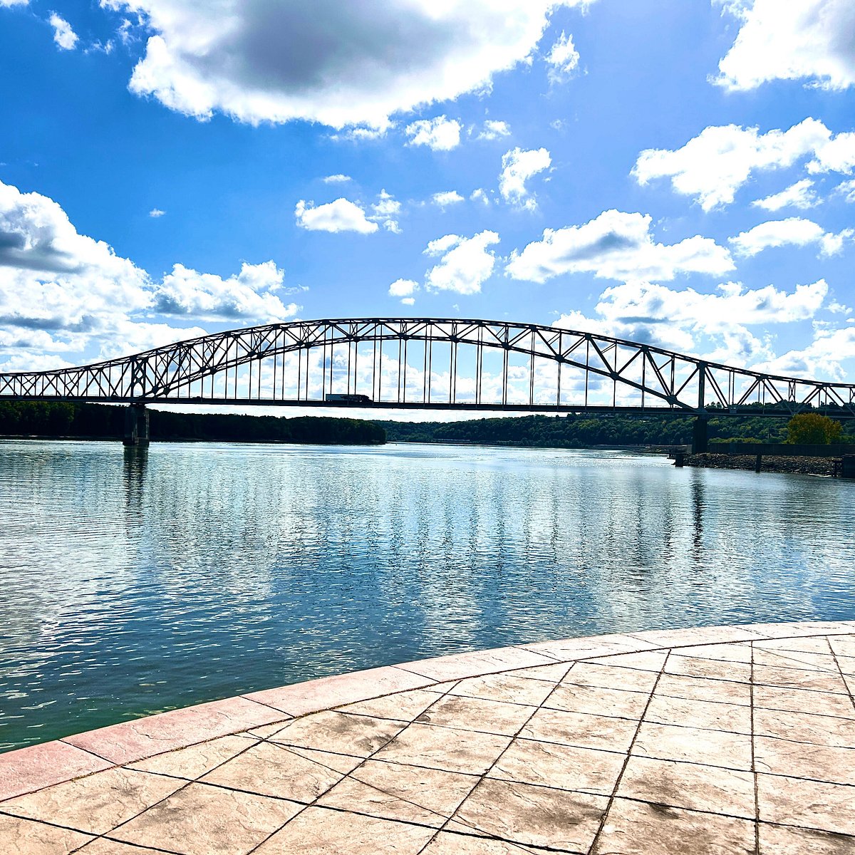 A wide, black steel truss bridge spans a calm, blue river under a bright sky with fluffy white clouds.
