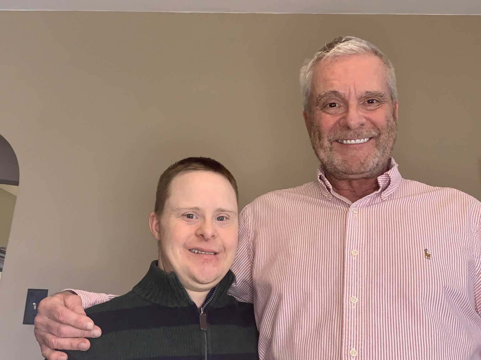 Two men, one with Down syndrome, smile as they pose for a photo, with the man on the right with his arm around the man on the left.