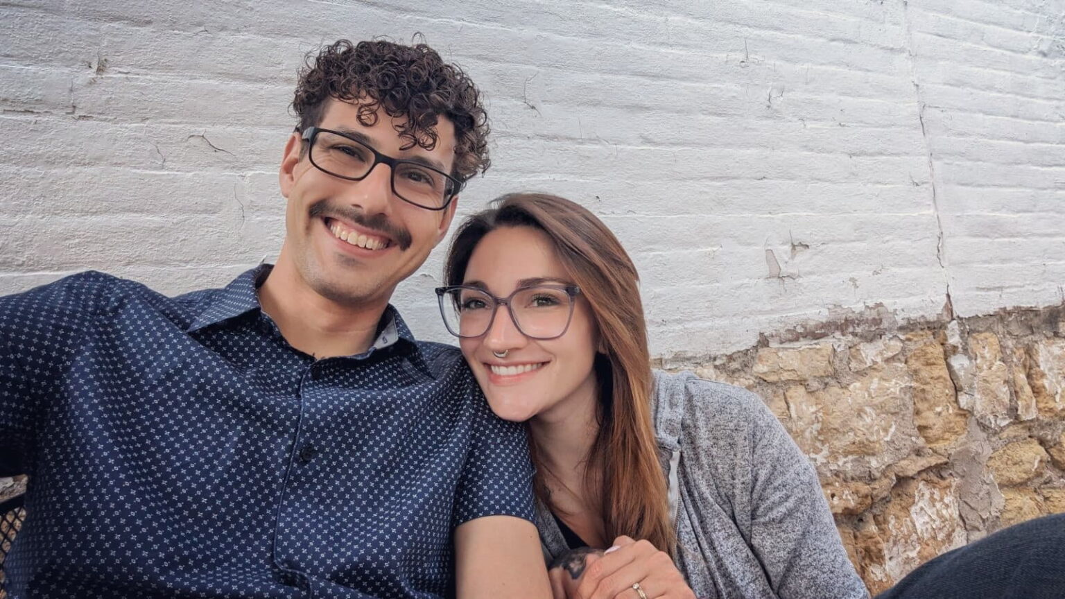 A smiling couple, a man with curly hair and glasses, and a woman with long brown hair and glasses, pose for a selfie against a white brick wall.