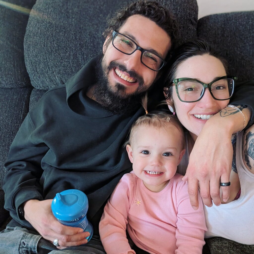 A happy family of three, a man, a woman, and a baby, are posing for a photo on a couch.