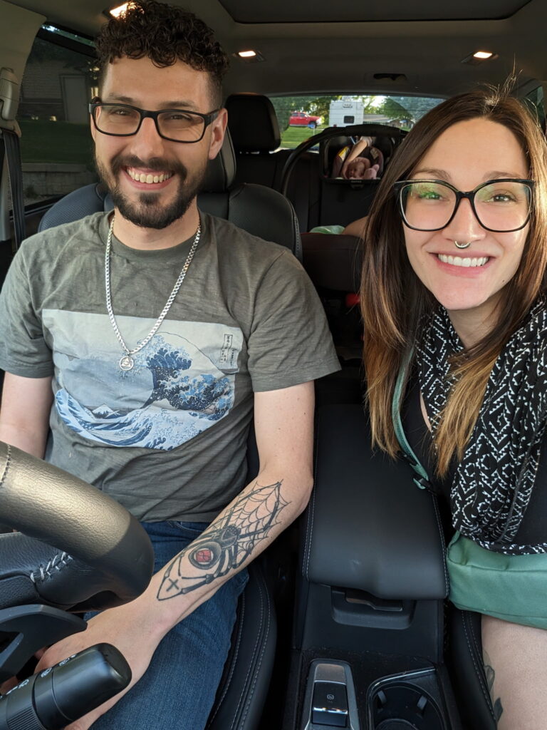 A smiling couple, a man with glasses and a beard and a woman with glasses and a nose ring, are in the front seats of a car, with a baby in a car seat visible in the back.