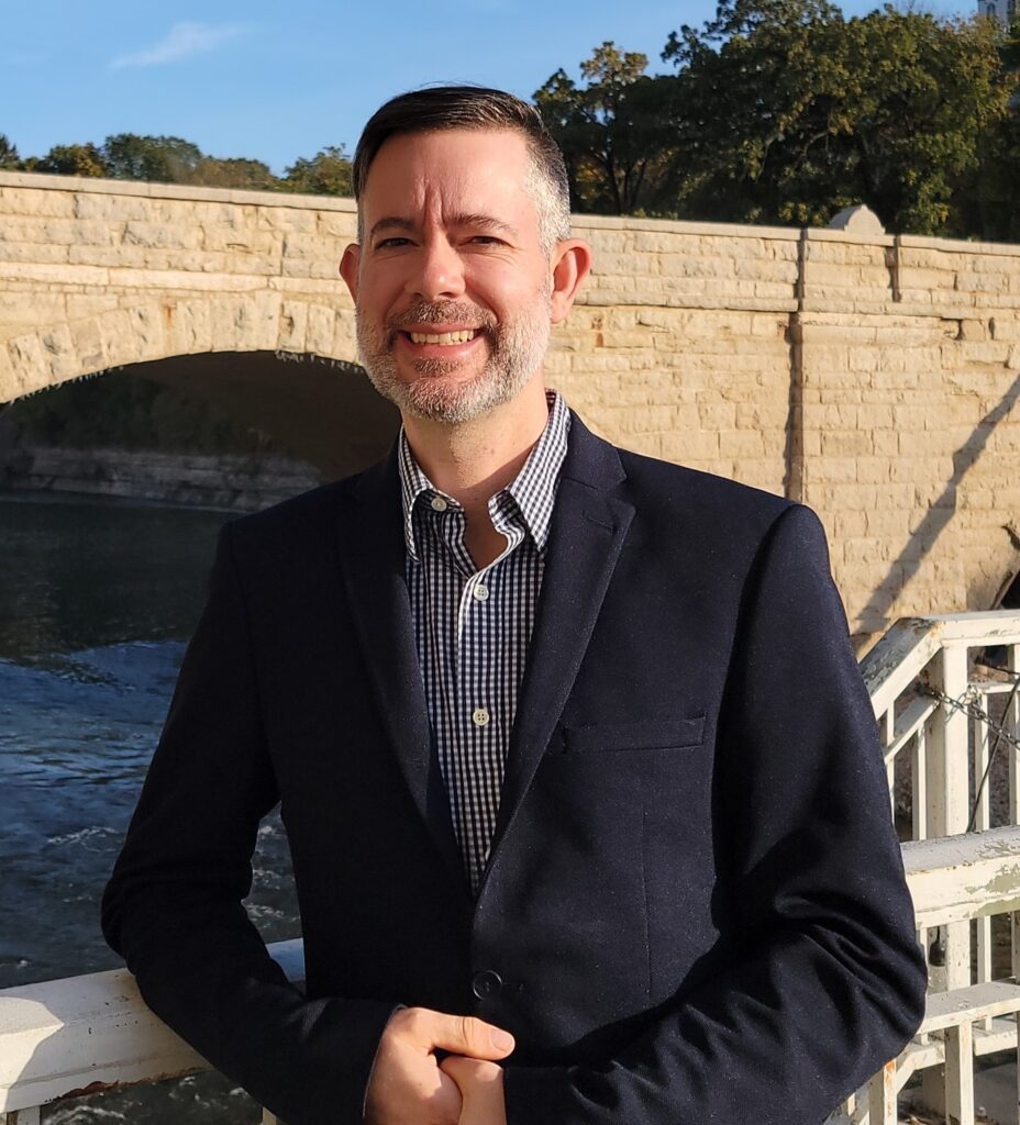 A man in a dark suit jacket and a checkered shirt smiles at the camera with a stone bridge and a body of water behind him.