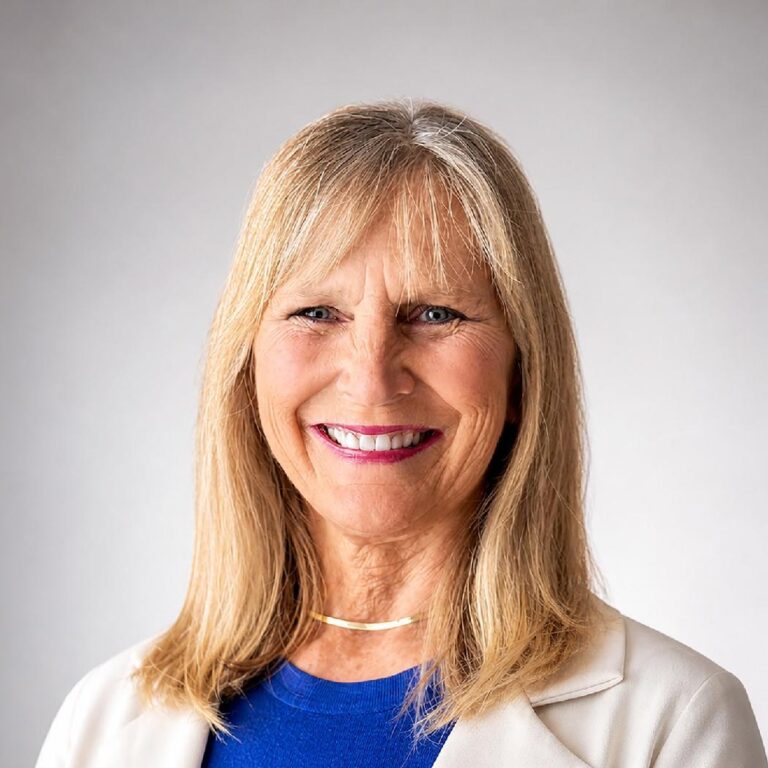 A headshot of a smiling, blonde woman wearing a blue shirt and a cream-colored blazer.