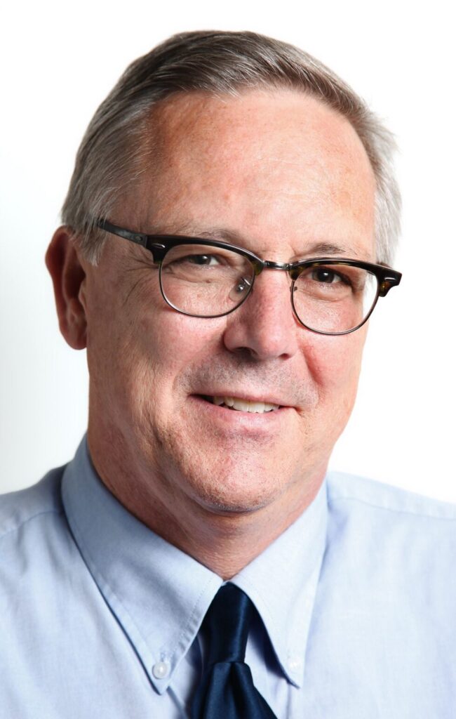 A close-up portrait of a middle-aged white man with graying hair, wearing glasses, a light blue collared shirt, and a dark blue tie.