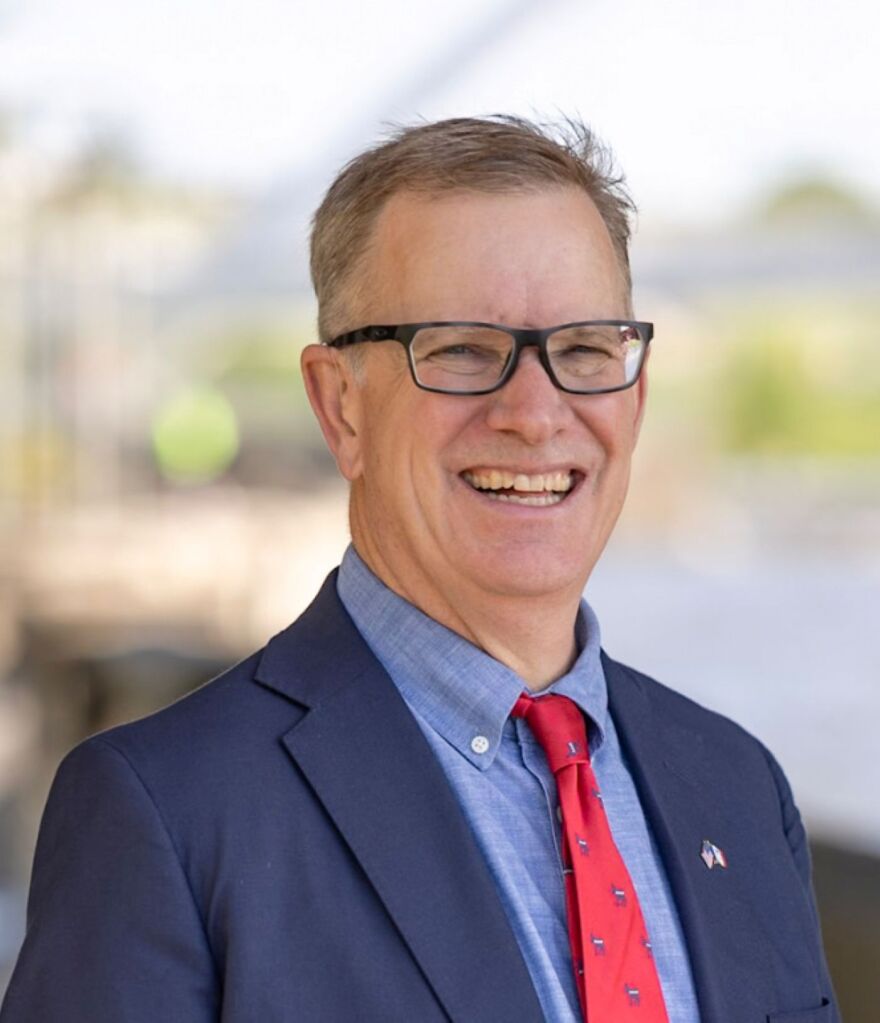 A smiling man wearing glasses, a blue suit, and a red tie.