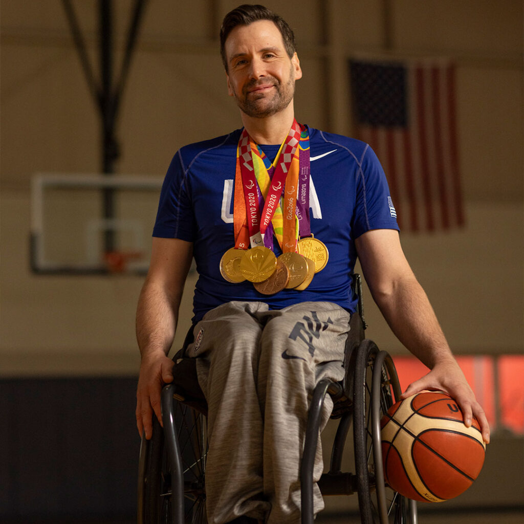 A smiling man in a wheelchair wearing a blue USA shirt with multiple gold and bronze medals around his neck, holding a basketball.