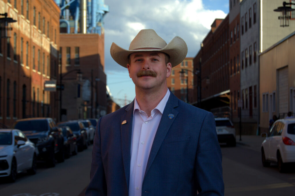 A man wearing a cowboy hat and a suit stands in the middle of a street lined with brick buildings and parked cars.