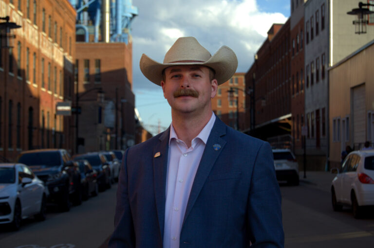 A man wearing a cowboy hat and a suit stands in the middle of a street lined with brick buildings and parked cars.