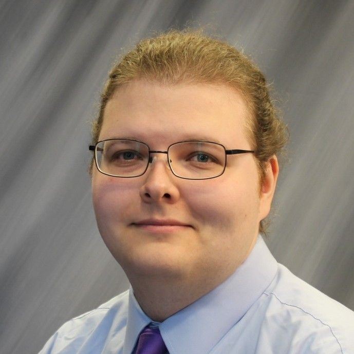 A headshot of a young man wearing glasses and a light blue dress shirt with a purple tie.