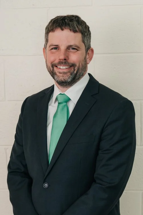 A man in a dark suit and green tie smiles at the camera.