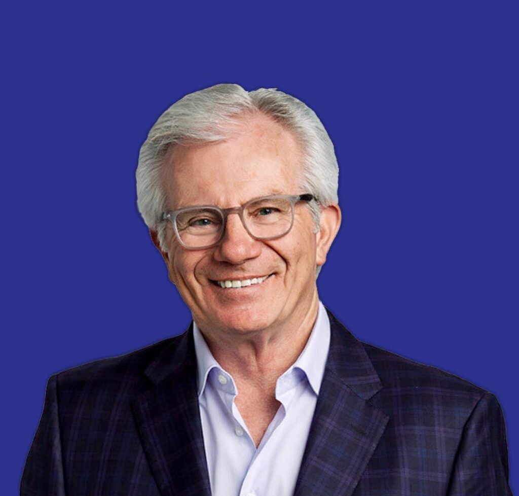 A smiling, older white man with gray hair and glasses wears a suit and a light blue shirt.