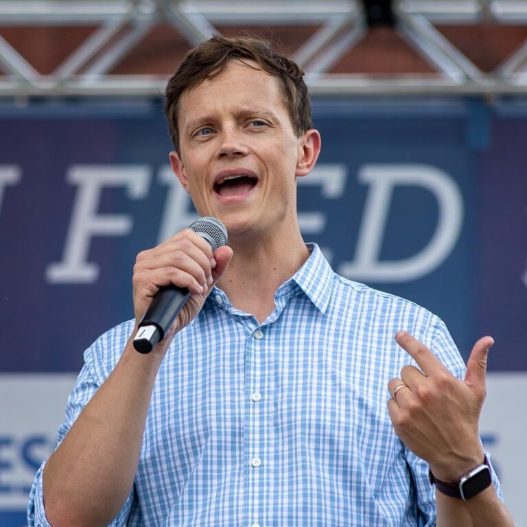 A man in a blue and white checkered shirt speaks into a microphone while gesturing with his right hand.