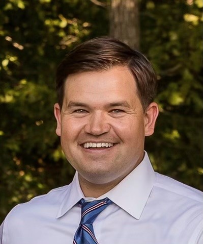 A man in a white collared shirt and a blue and red striped tie smiles broadly, with trees in the background.