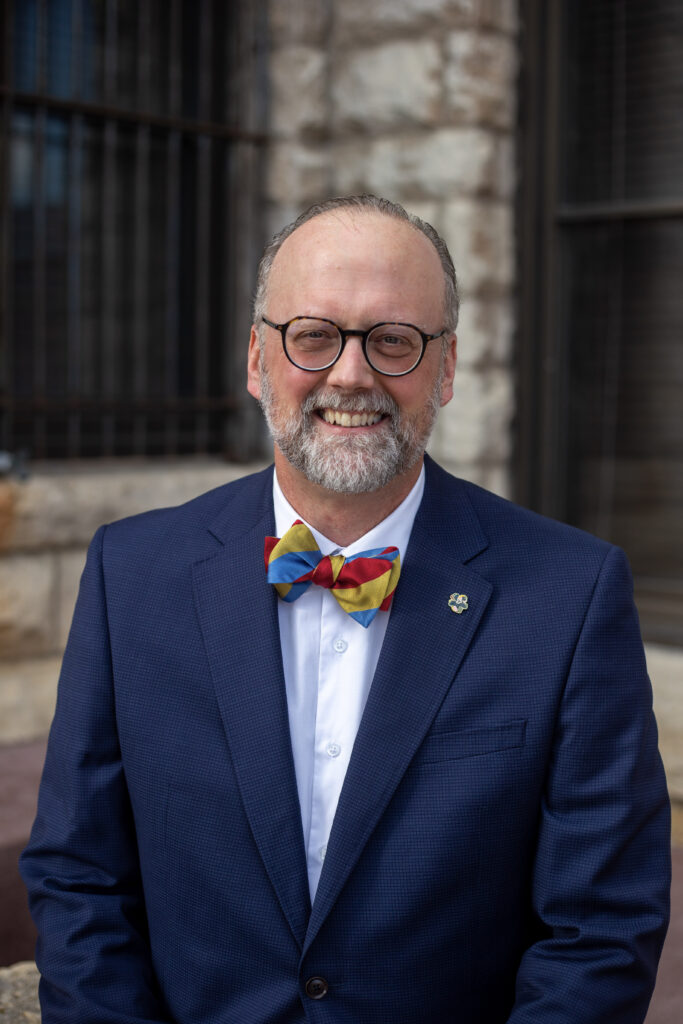 A smiling man with a beard and glasses wears a blue suit, a white shirt, and a colorful bow tie.