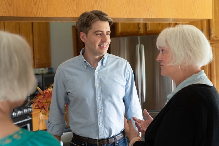 A young man in a light blue button-down shirt smiles and talks to an older woman with white hair, who is wearing a black cardigan.