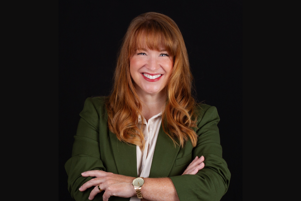 A woman with red hair and a green blazer smiles with her arms crossed.