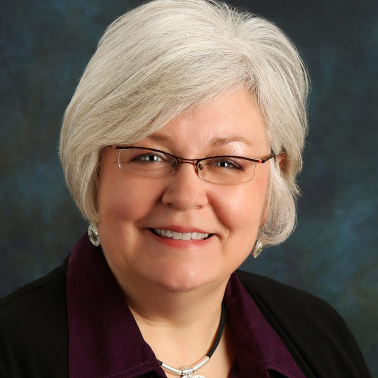 A headshot of a middle-aged woman with short, gray hair, wearing glasses, a black jacket over a purple shirt, and a large silver pendant necklace.