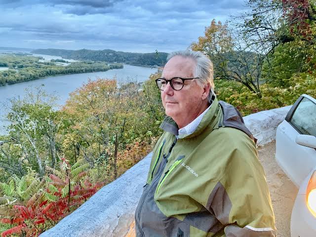 Man in a green jacket and glasses stands on a ledge overlooking a river and trees with fall foliage.