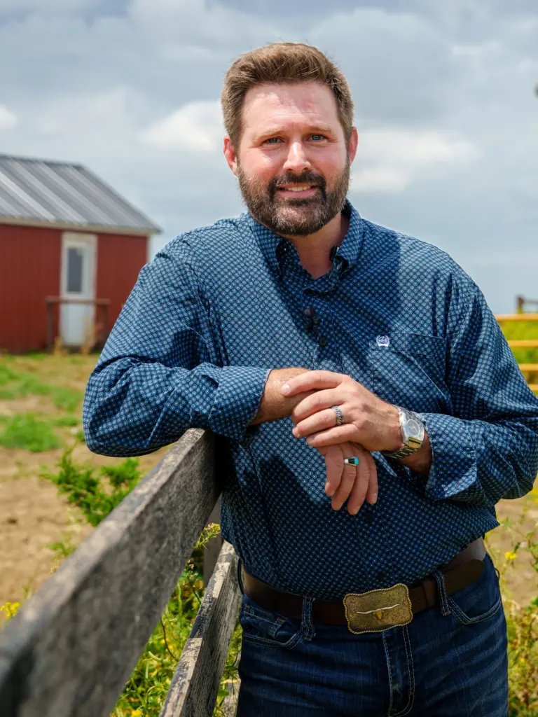 A man with a beard and blue eyes, wearing a blue patterned shirt, jeans, and a belt with a large buckle, leans on a wooden fence in front of a red barn.