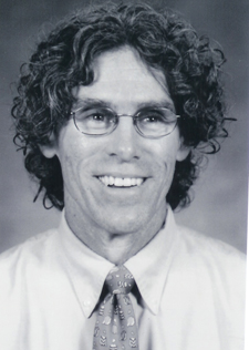 Black and white headshot of a man with curly hair and glasses, smiling.