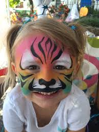 A young girl with blonde pigtails smiles, her face painted like a tiger with pink, orange, black, and white stripes and markings.