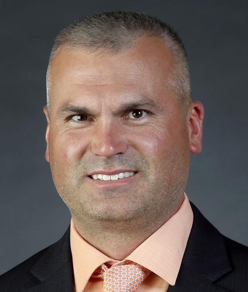 A headshot of a man with short hair, wearing a dark suit, an orange collared shirt, and an orange tie.