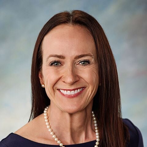 A woman with brown hair, wearing a navy blue dress and a pearl necklace, smiles at the camera.