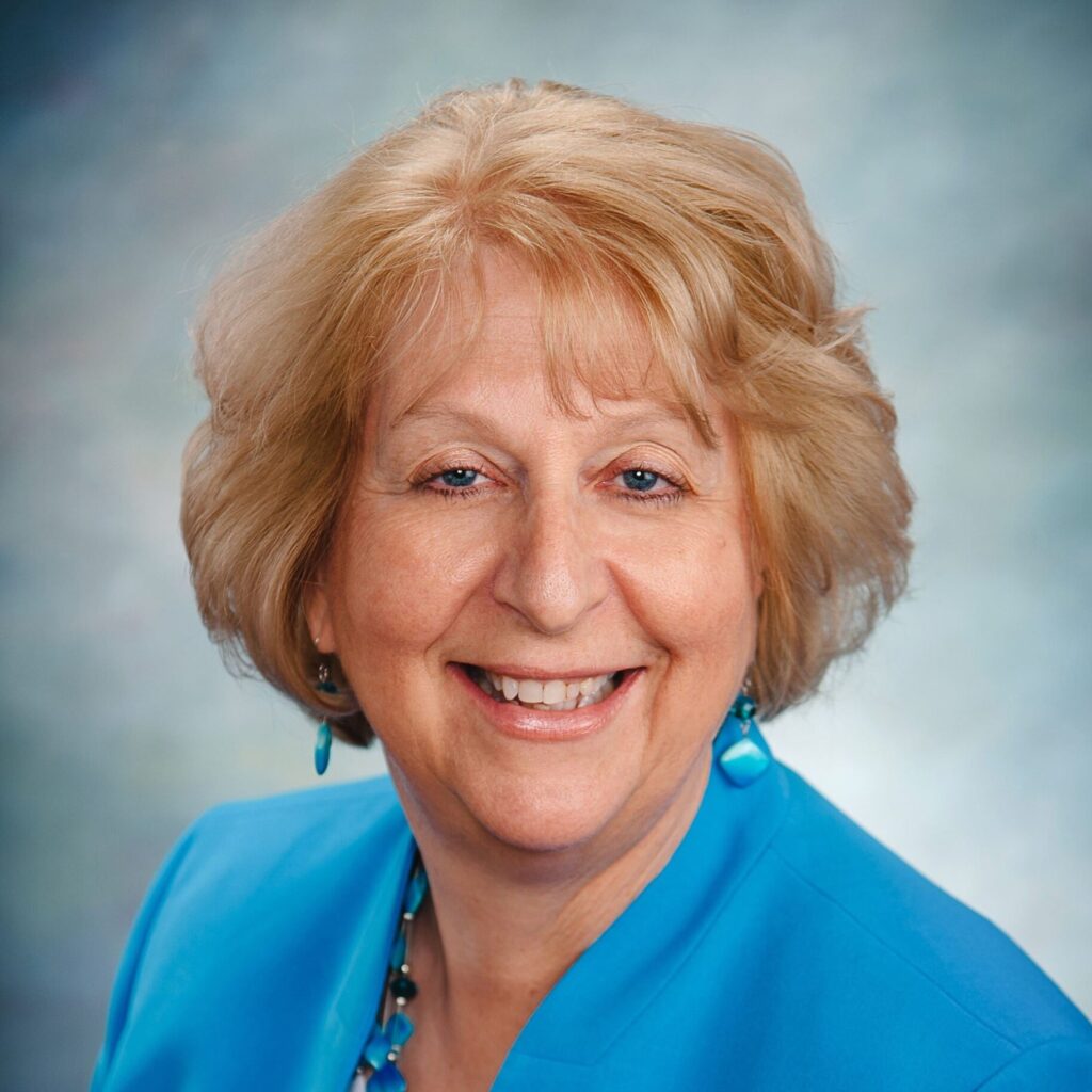 A headshot of an older woman with blonde hair and blue eyes, smiling and wearing a blue jacket and necklace.