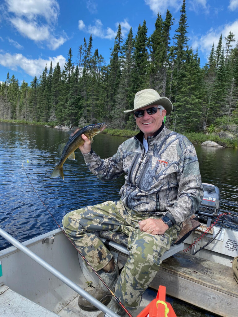 A smiling man in camouflage clothing and a hat holds up a fish while sitting in a boat on a lake, with a forest and blue sky in the background.