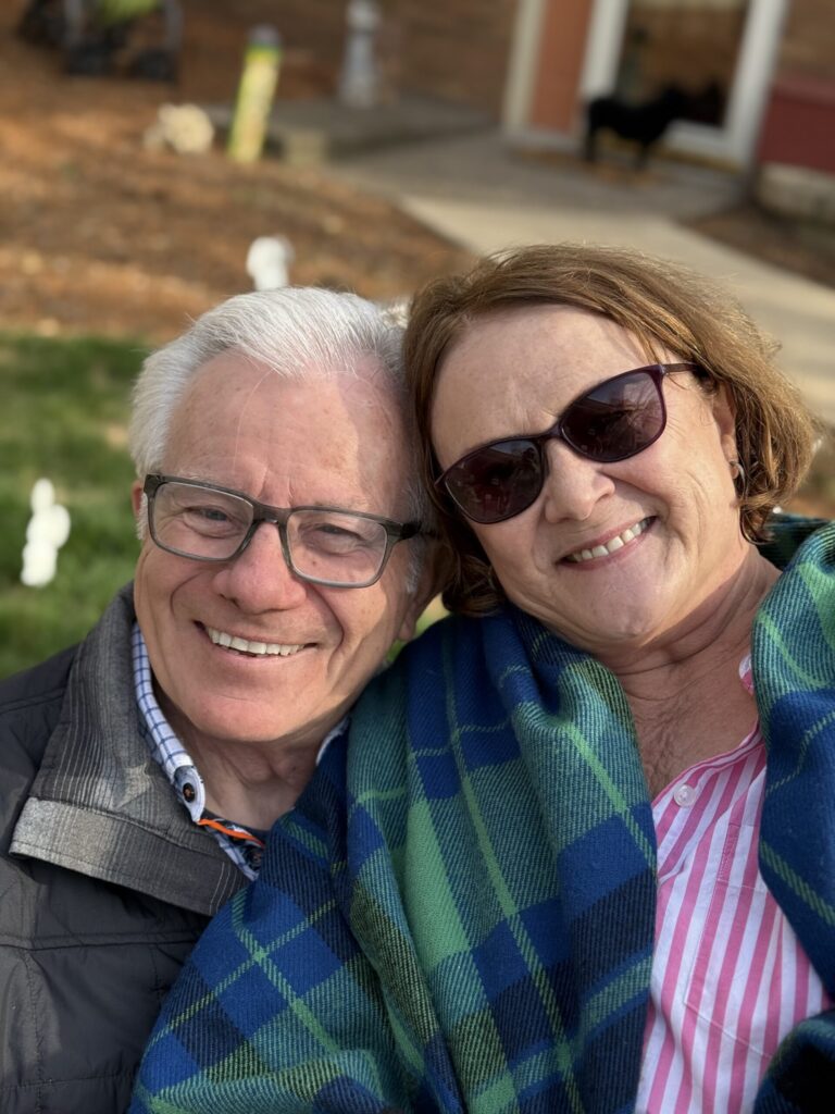 Rick Dickinson and his wife, Rae Ann, smile together outdoors while wrapped in a blue and green plaid blanket.