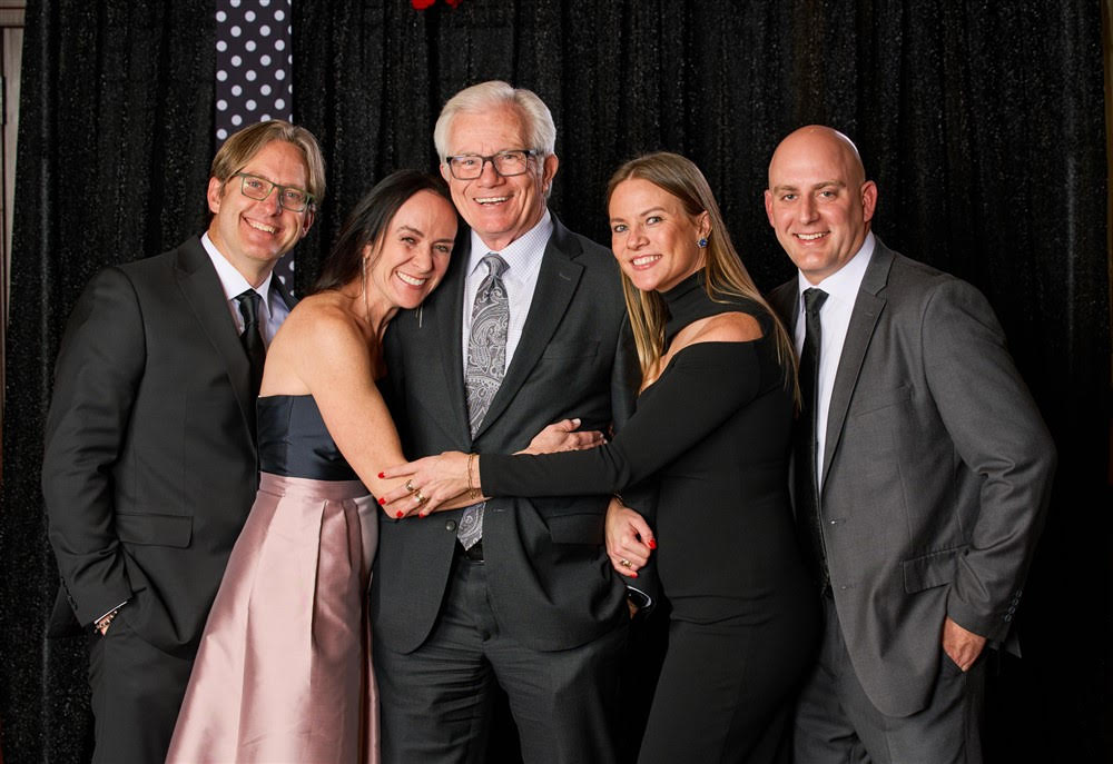 A group of five people, three men and two women, pose for a photo in formal attire against a black backdrop with a polka dot pattern.