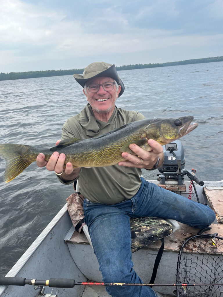 A smiling man in a hat and glasses sits in a boat holding a large walleye fish, with a Yamaha outboard motor visible behind him.