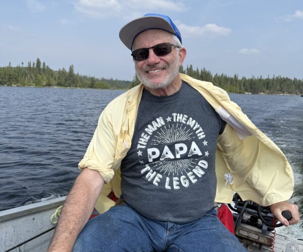 A smiling man wearing a blue baseball cap, sunglasses, and a t-shirt that reads "The Man, The Myth, Papa, The Legend" sits in a boat on a lake.