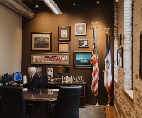 Rick Dickinson works at a desk in an office with American and Iowa flags, framed photographs, and Dubuque memorabilia.