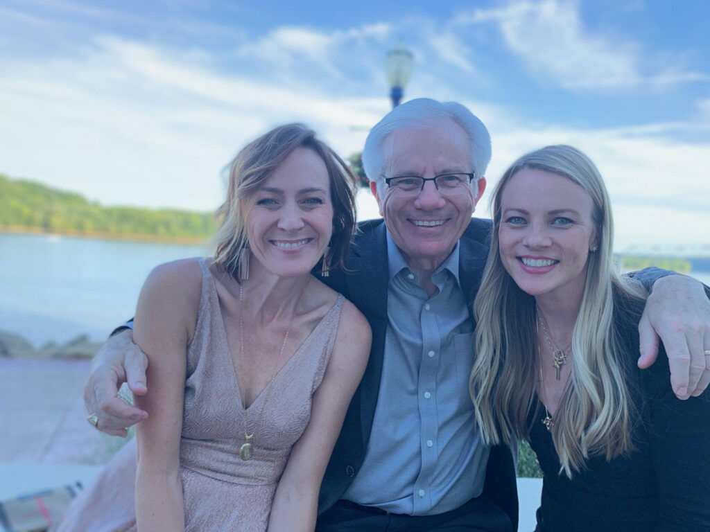A man with white hair and glasses smiles between two women, one on his left and one on his right, all posing for a photo outdoors.
