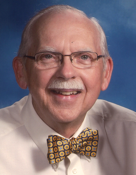 A smiling, balding white man with glasses and a mustache wears a white shirt and a patterned bow tie.