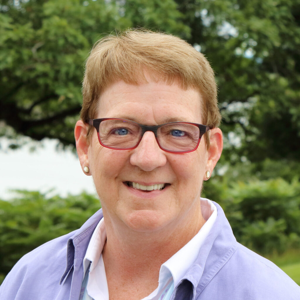 A smiling woman with short blonde hair and blue eyes, wearing glasses and a light purple shirt over a white blouse, poses outdoors with a blurred green background.