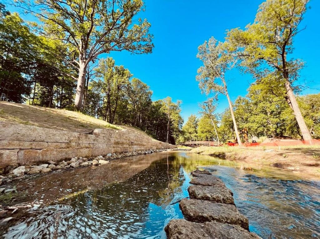 A serene river flows through a park lined with trees and a stone wall, with a bridge visible in the distance.