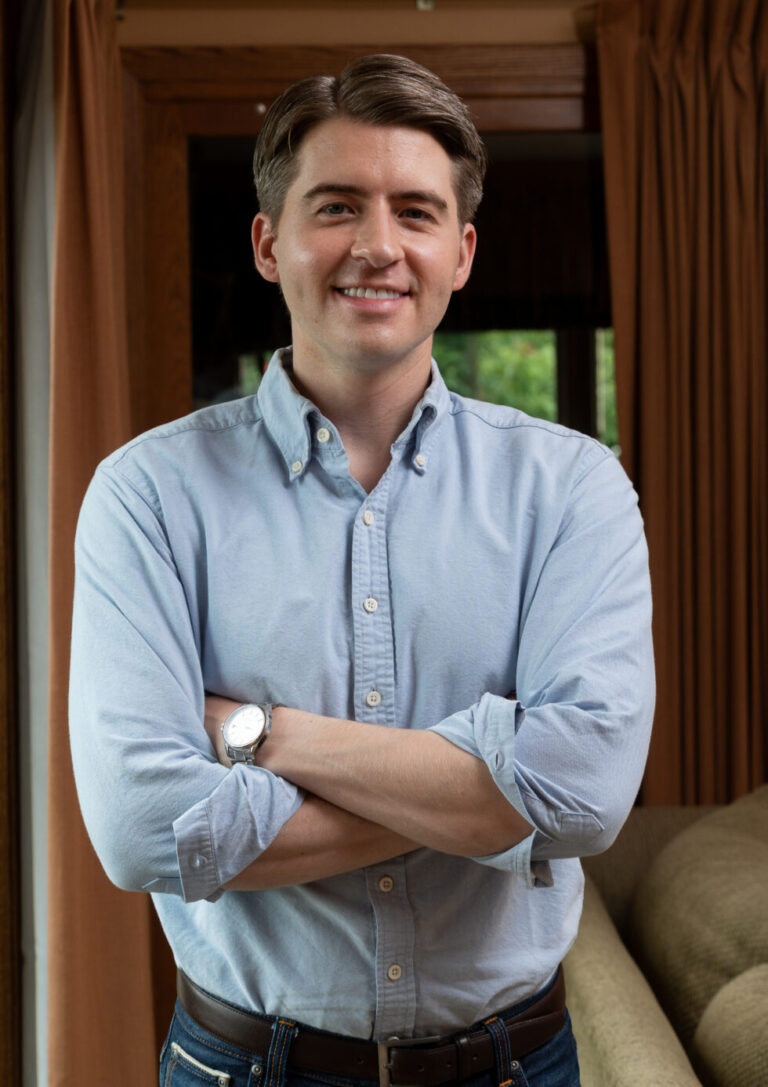 A man in a light blue button-down shirt and jeans smiles with his arms crossed.